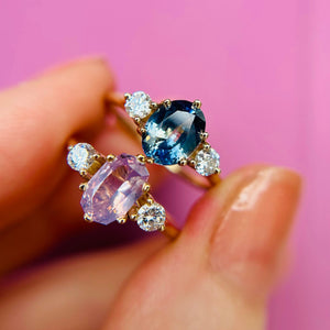 Two elegant engagement rings - one deep blue oval like the sea and one pretty lilac -  with diamond accent stones either side of the centre gem, held in a person's fingers against a pink background.