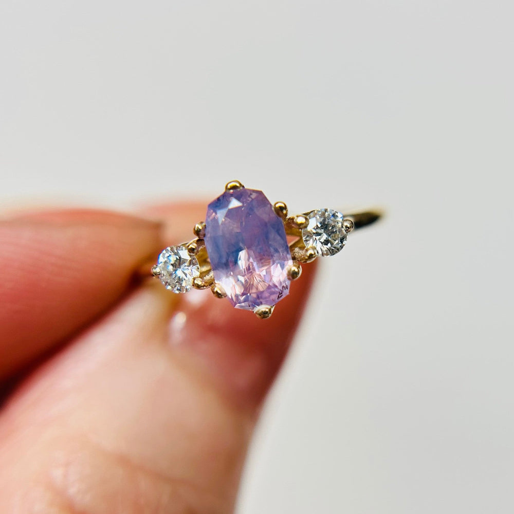 A lilac and purple sapphire 3 stone engagement ring, with two round diamond accent stones on either side. Held inbetween two fingers.