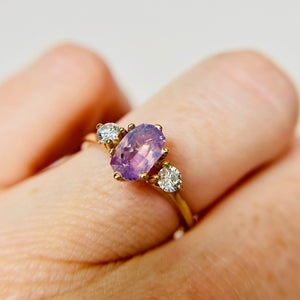 A lilac oval engagement ring set in yellow gold is worn on a finger.