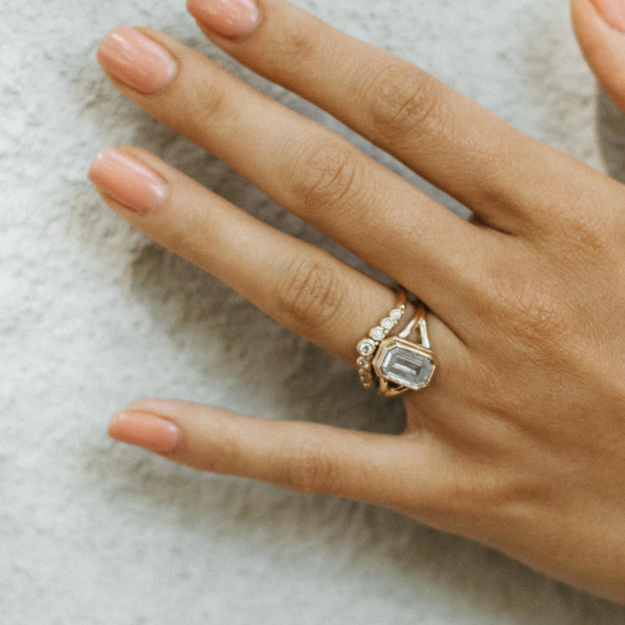 A yellow gold ring with a cushion-cut salt and pepper diamond at it's centre. It has a modern split shank setting and is displayed on a reflective surface with a soft, textured background.