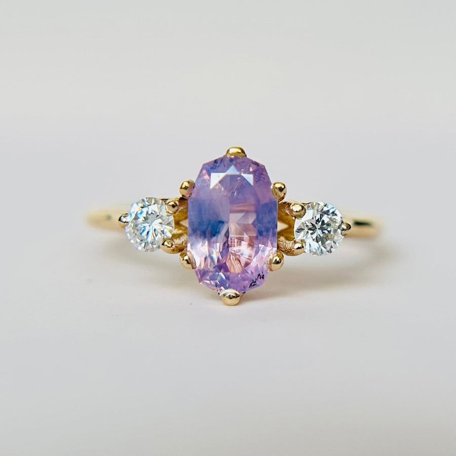 A lilac and purple sapphire 3 stone engagement ring, with two round diamond accent stones on either side. Held inbetween two fingers.