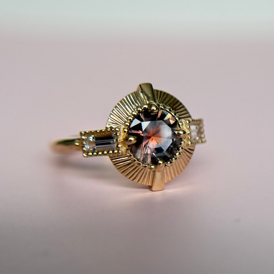Gold ring with a round pink sapphire gemstone held between fingers against a pink background