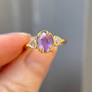 Gold engagement ring with a purple pink sapphire held by a hand against a neutral background