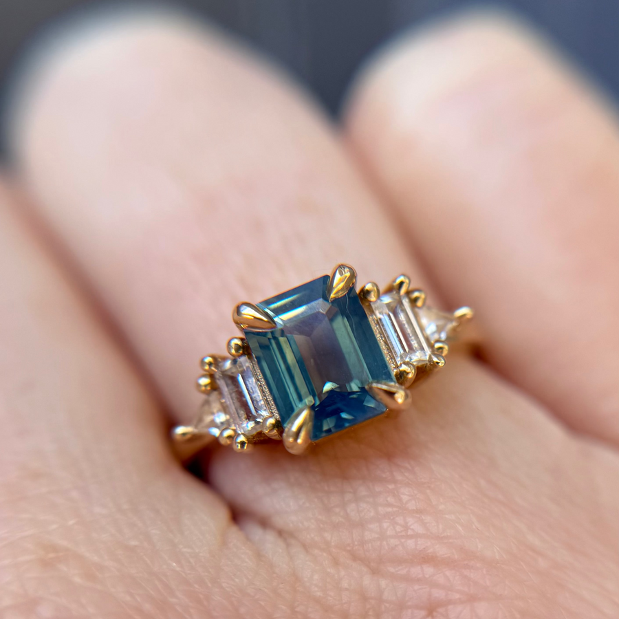 Yellow Gold engagement ring handmade in Toronto Canada with a blue and teal sapphire gemstone held by a hand against a pink background