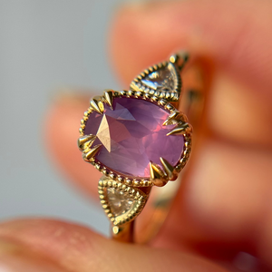 A Yellow Gold engagement ring with a purple and pink oval sapphire held between fingers against a blurred background.