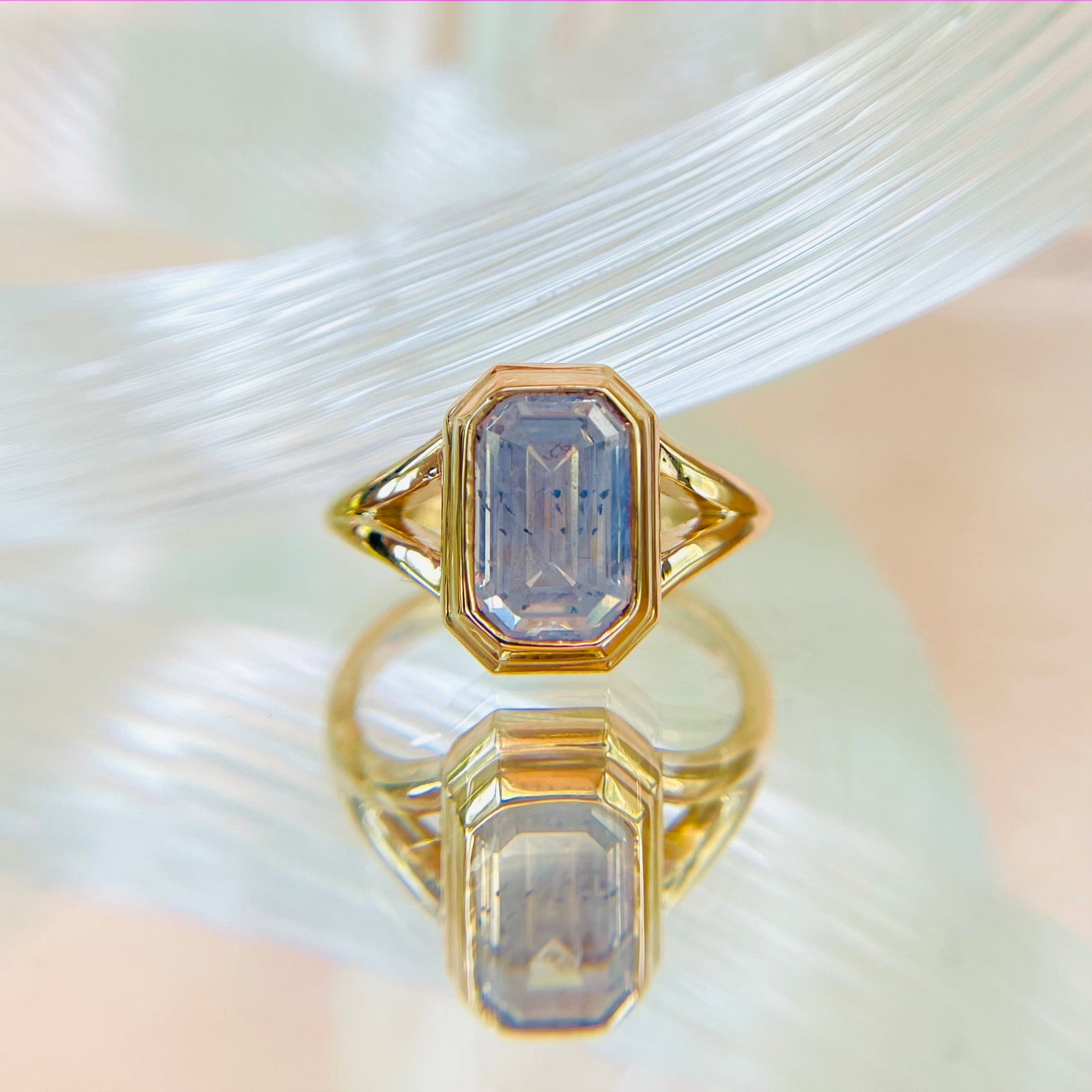 A yellow gold ring with a cushion-cut salt and pepper diamond at it's centre. It has a modern split shank setting and is displayed on a reflective surface with a soft, textured background.