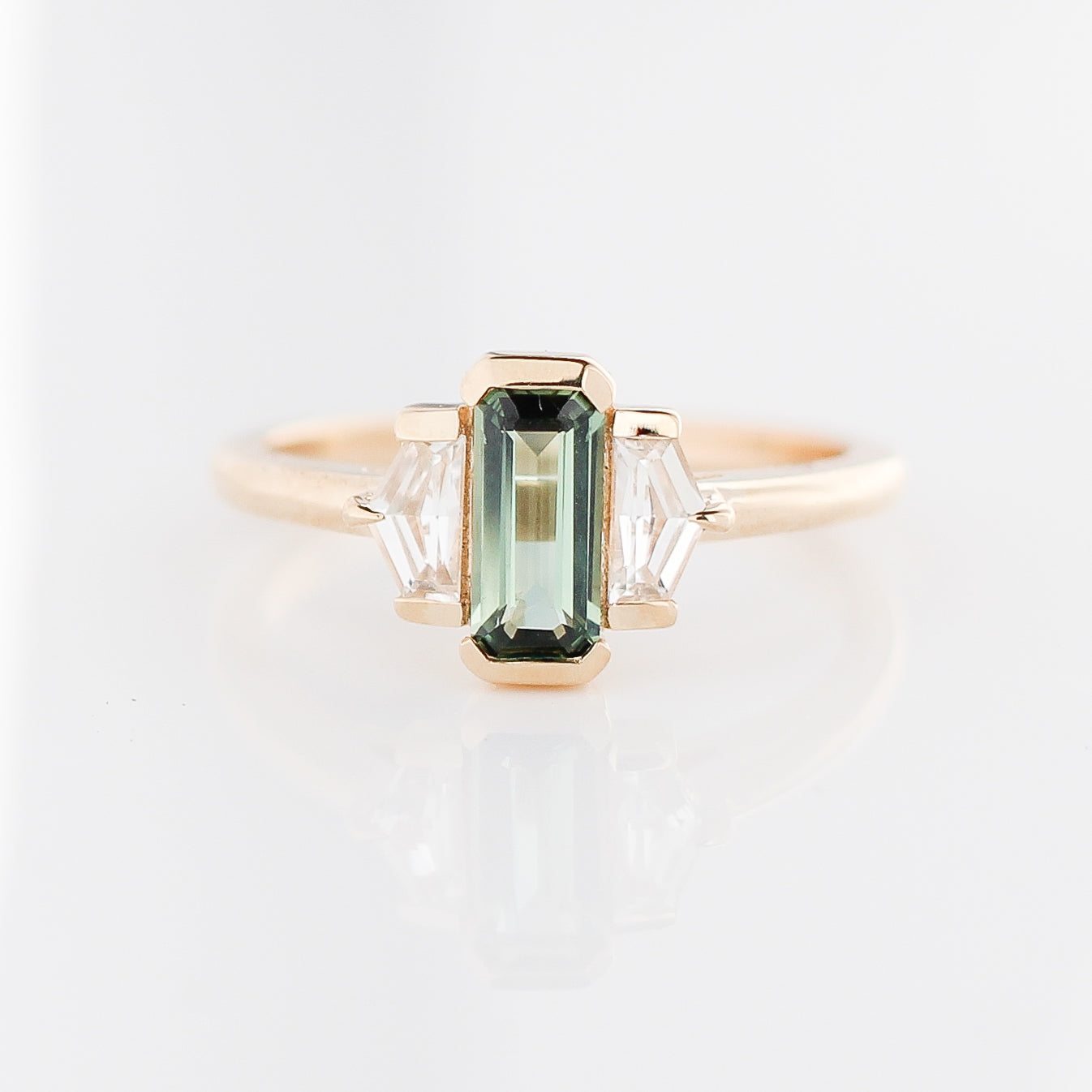 Three stone yellow gold ring, with elongated emerald cut green sapphire half bezel set and shield shaped white sapphire side stones in half bezel settings, front view