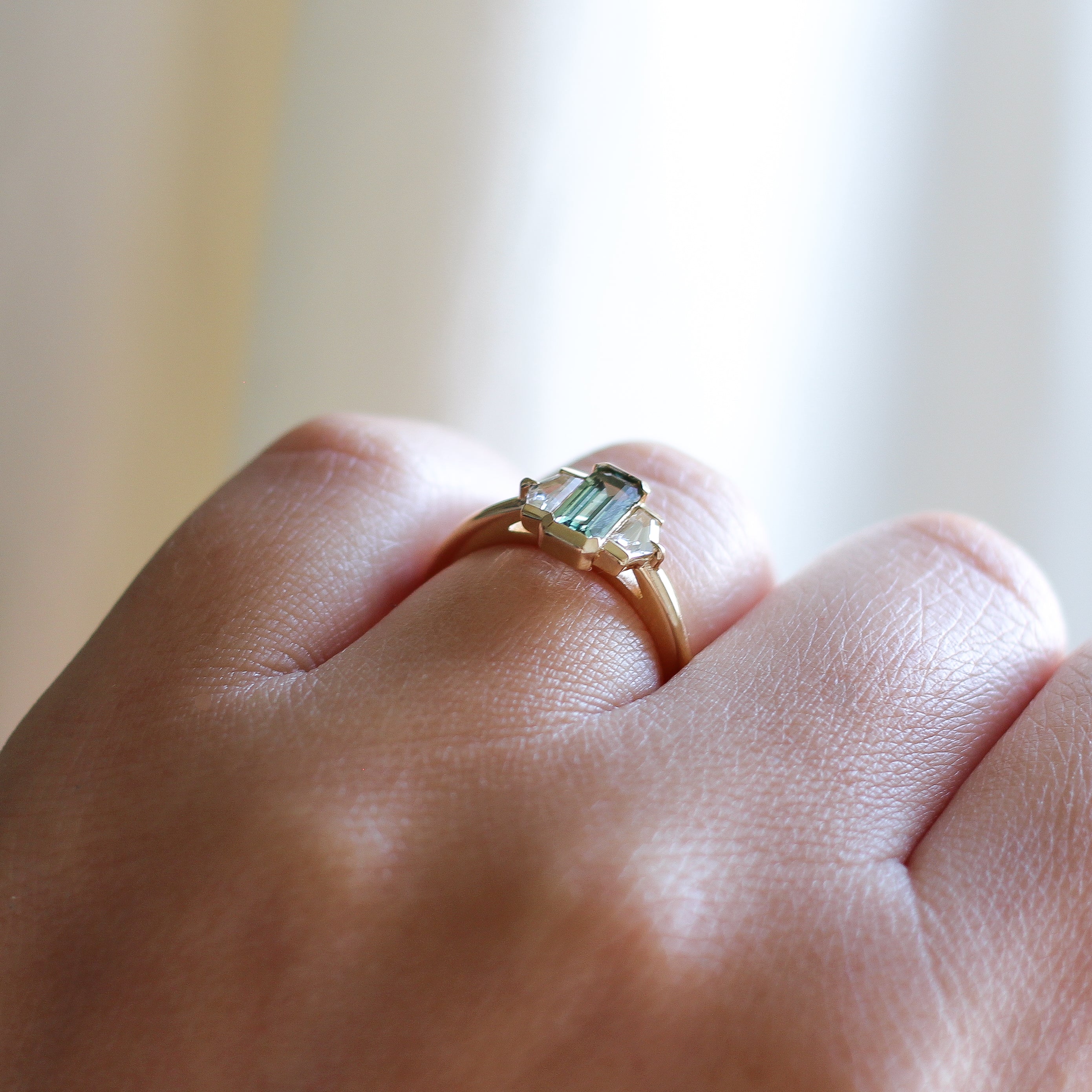 Three stone yellow gold ring, with elongated emerald cut green sapphire half bezel set and shield shaped white sapphire side stones in half bezel settings on hand, side view