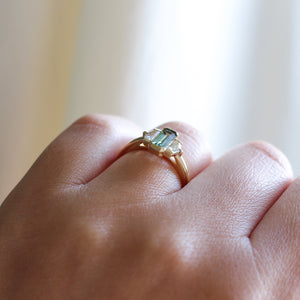 Three stone yellow gold ring, with elongated emerald cut green sapphire half bezel set and shield shaped white sapphire side stones in half bezel settings on hand, side view