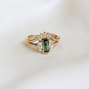 Three stone yellow gold ring, with elongated emerald cut green sapphire half bezel set and shield shaped white sapphire side stones in half bezel settings stacked with a yellow gold curved white diamond wedding band, front view