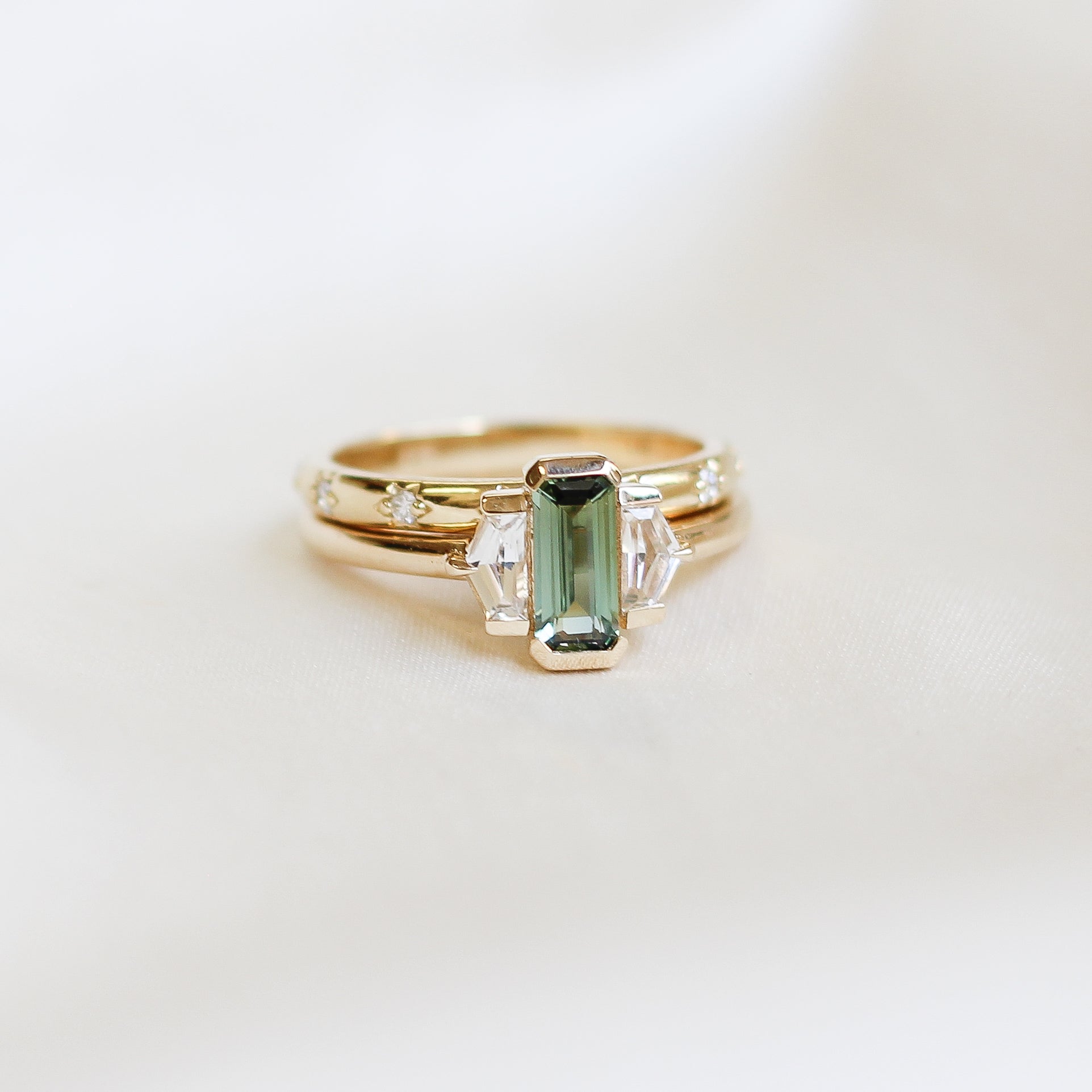 Three stone yellow gold ring, with elongated emerald cut green sapphire half bezel set and shield shaped white sapphire side stones in half bezel settings stacked with a yellow gold straight white diamond wedding band, front view