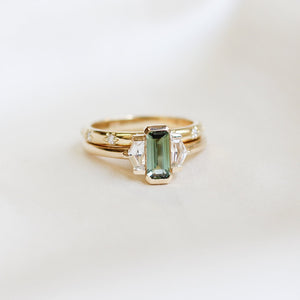 Three stone yellow gold ring, with elongated emerald cut green sapphire half bezel set and shield shaped white sapphire side stones in half bezel settings stacked with a yellow gold straight white diamond wedding band, front view