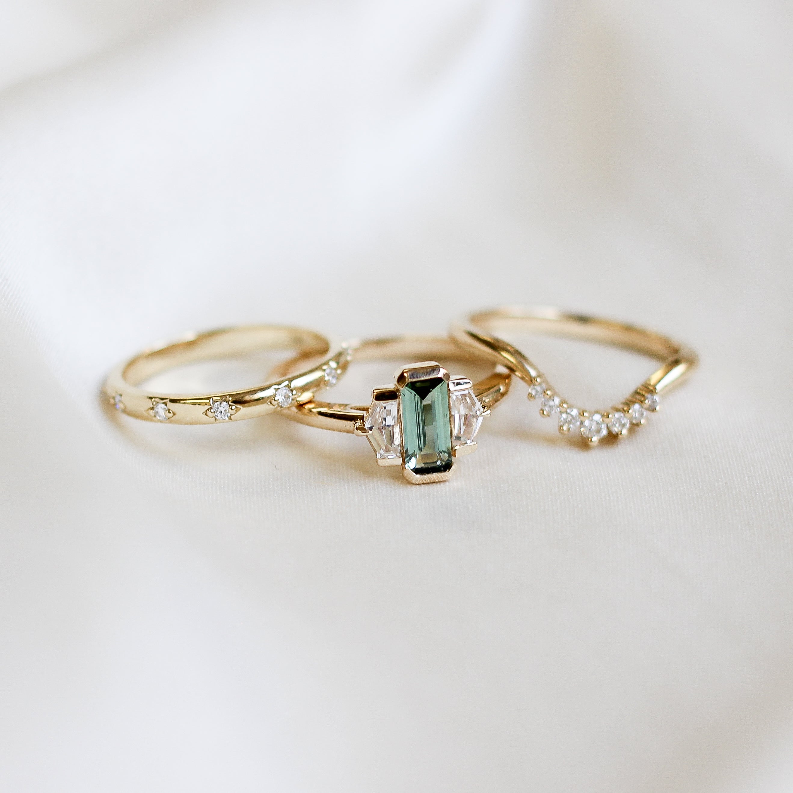 Three stone yellow gold ring, with elongated emerald cut green sapphire half bezel set and shield shaped white sapphire side stones in half bezel settings in between two yellow gold white diamond wedding bands, front view