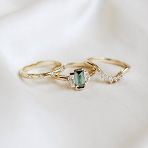 Three stone yellow gold ring, with elongated emerald cut green sapphire half bezel set and shield shaped white sapphire side stones in half bezel settings in between two yellow gold white diamond wedding bands, front view