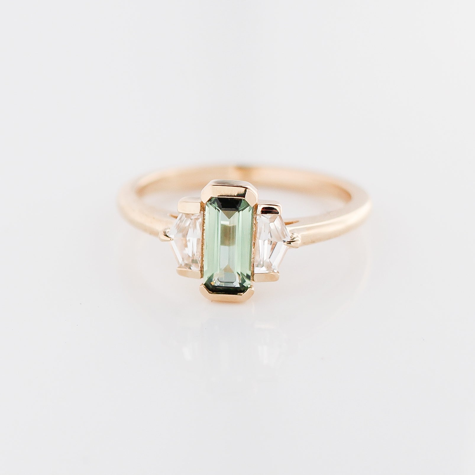Three stone yellow gold ring, with elongated emerald cut green sapphire half bezel set and shield shaped white sapphire side stones in half bezel settings, front view