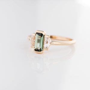 Three stone yellow gold ring, with elongated emerald cut green sapphire half bezel set and shield shaped white sapphire side stones in half bezel settings, quarter view
