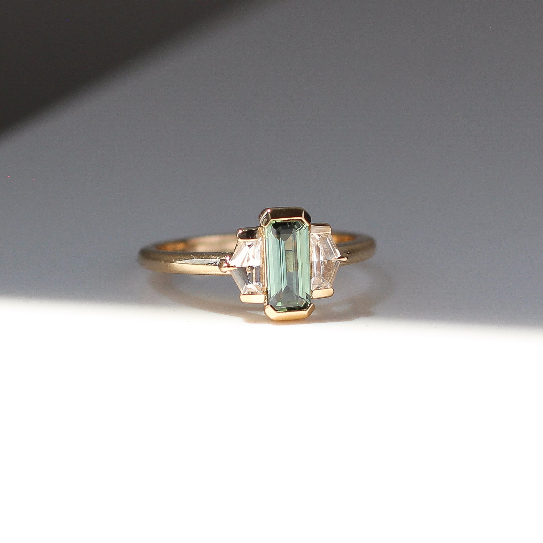 Three stone yellow gold ring, with elongated emerald cut green sapphire half bezel set and shield shaped white sapphire side stones in half bezel settings, quarter view