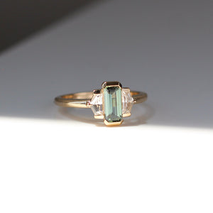 Three stone yellow gold ring, with elongated emerald cut green sapphire half bezel set and shield shaped white sapphire side stones in half bezel settings, quarter view