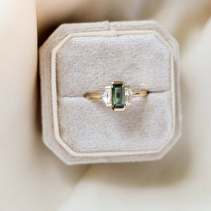 Three stone yellow gold ring, with elongated emerald cut green sapphire half bezel set and shield shaped white sapphire side stones in half bezel settings in ring box, top view
