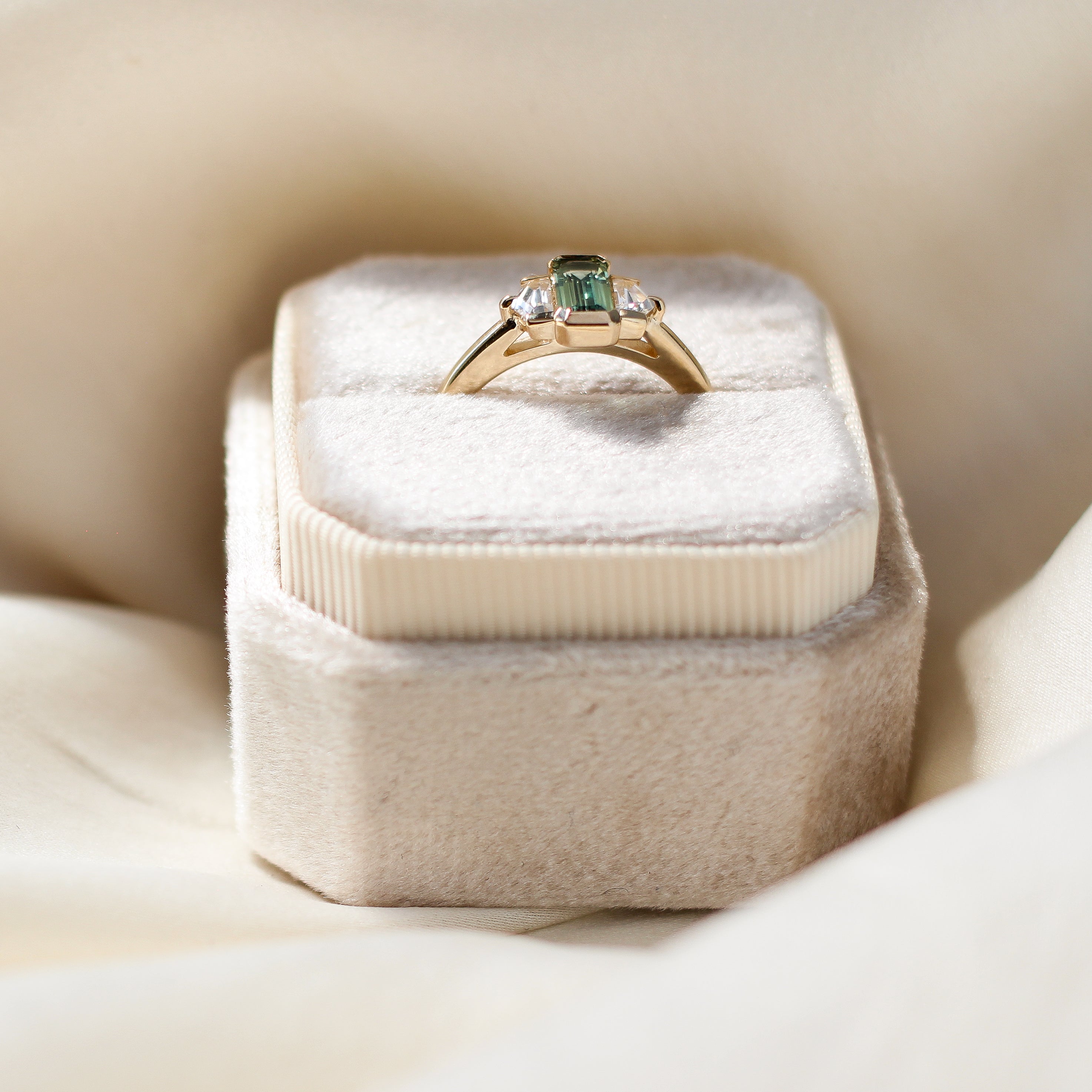Three stone yellow gold ring, with elongated emerald cut green sapphire half bezel set and shield shaped white sapphire side stones in half bezel settings in ring box, side view
