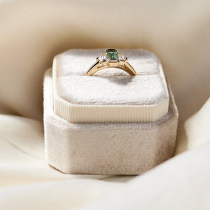 Three stone yellow gold ring, with elongated emerald cut green sapphire half bezel set and shield shaped white sapphire side stones in half bezel settings in ring box, side view