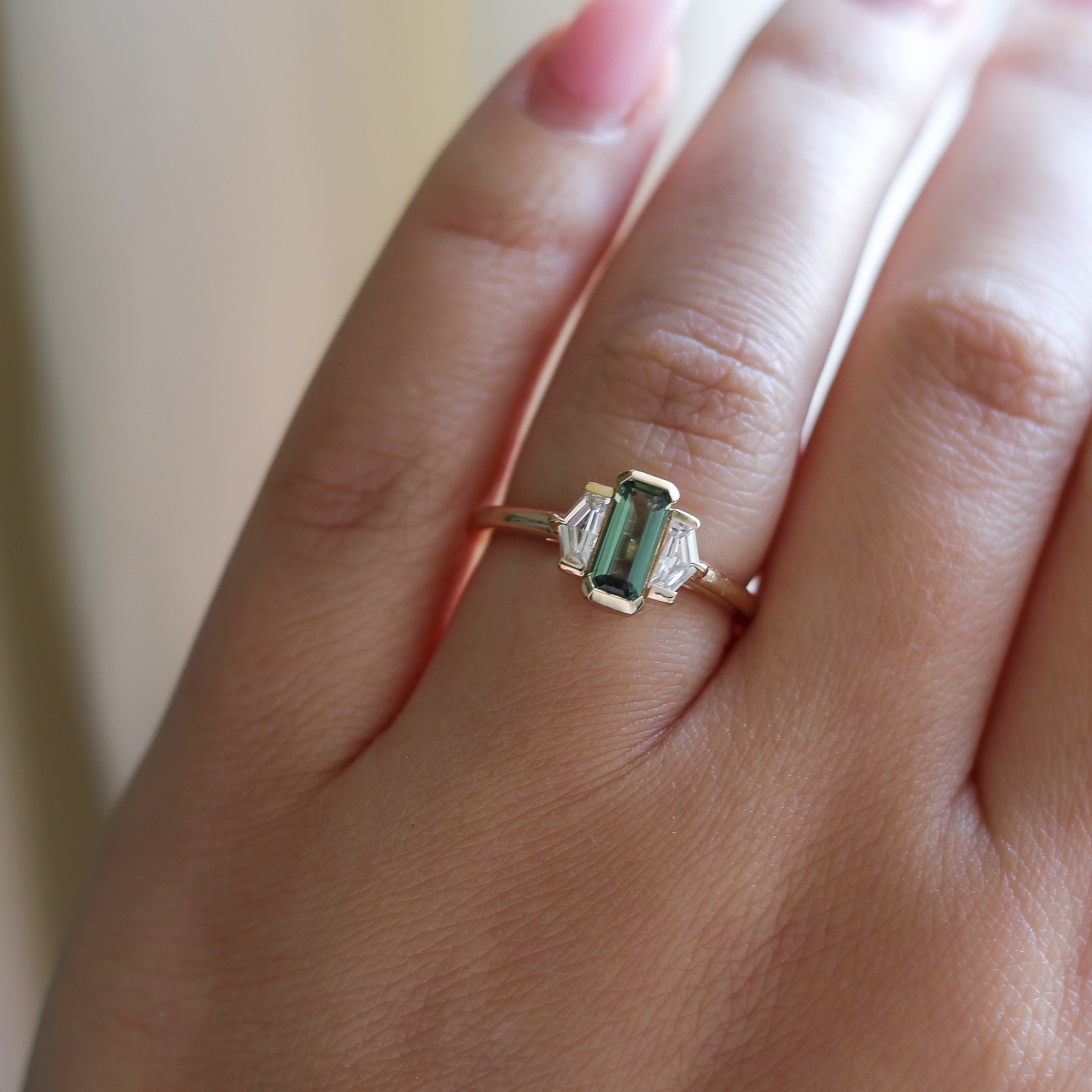 Three stone yellow gold ring, with elongated emerald cut green sapphire half bezel set and shield shaped white sapphire side stones in half bezel settings on hand, top view