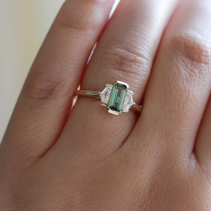 Three stone yellow gold ring, with elongated emerald cut green sapphire half bezel set and shield shaped white sapphire side stones in half bezel settings on hand, top view
