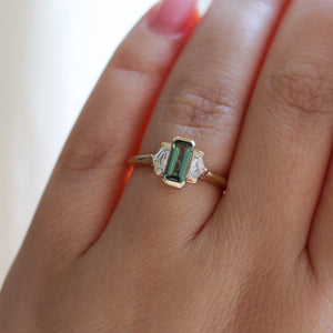 Three stone yellow gold ring, with elongated emerald cut green sapphire half bezel set and shield shaped white sapphire side stones in half bezel settings on hand, top view