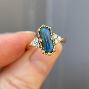 Gold engagement ring with a blue hexagon shaped sapphire and sparkling diamond accents held by a hand against a neutral background