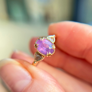 3 stone engagement ring with a bright purple almost pink sapphire gemstone, with diamond accents on the band held between fingers against a blurred background