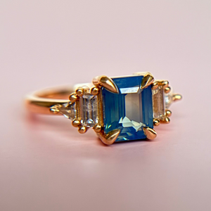 Yellow Gold 3-stone engagement ring reminiscent of an art-deco style with a blue sapphire on a soft pink background. 