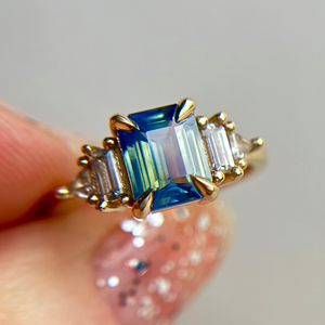 Yellow Gold ring with a Teal blue center sapphire gemstone held by a hand against a neutral background