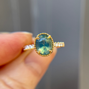 Gold engagement ring with a teal green sapphire held between fingers against a neutral background