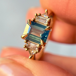Yellow Gold engagement ring with blue sapphire center gemstone and sparkling white accent stones held between fingers against a neutral background.