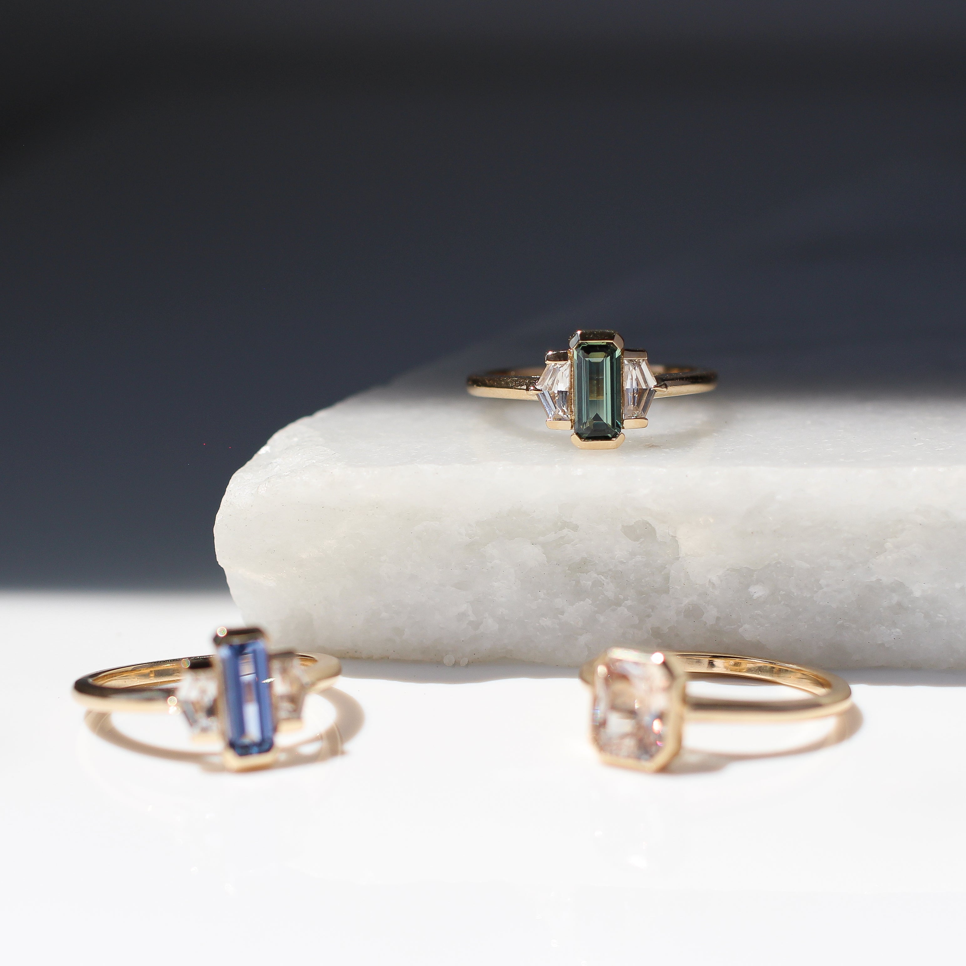 Three yellow gold engagement rings with coloured sapphires on a marble surface