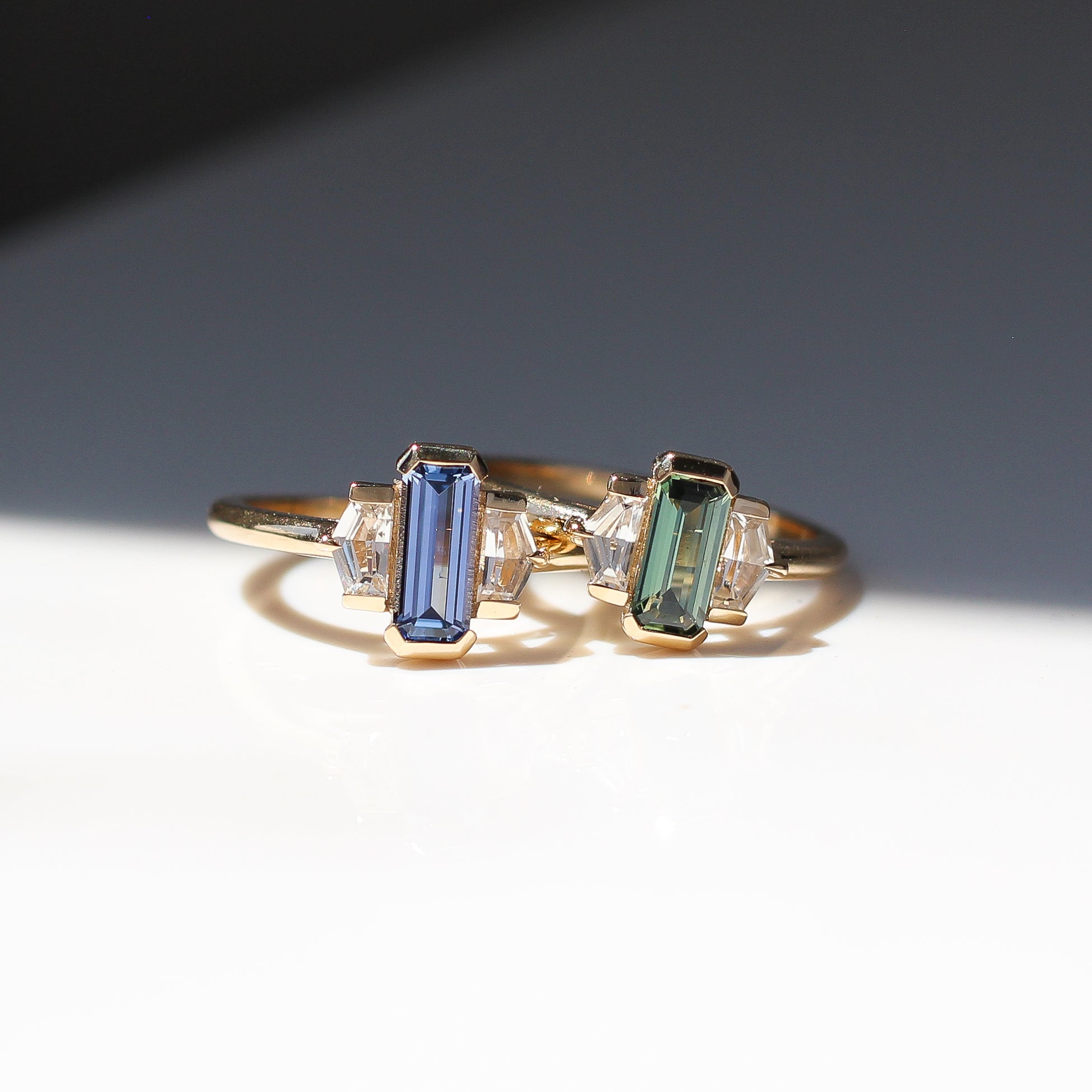 Two yellow gold engagement rings with half bezel set coloured sapphires and white sapphire shield accents side by side, front view