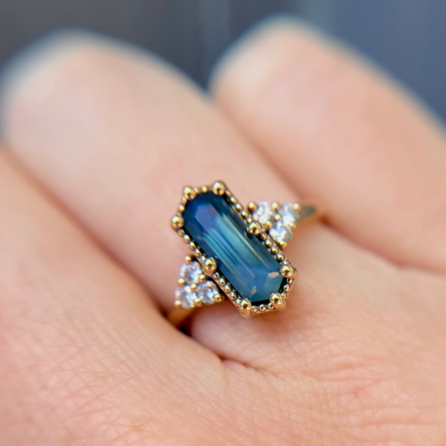 Gold ring with a blue parti or bi-color sapphire and diamond accent stones held between fingers against a blurred background