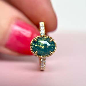 Engagement Ring made in Canada with a teal and green centre gemstone and sparkling diamond accents held by a hand with pink nail polish on a blurred background